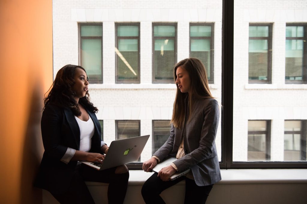 woman-holding-laptop-computer-near-window-1181563 Two women having a business discussion in an office setting, using a laptop.